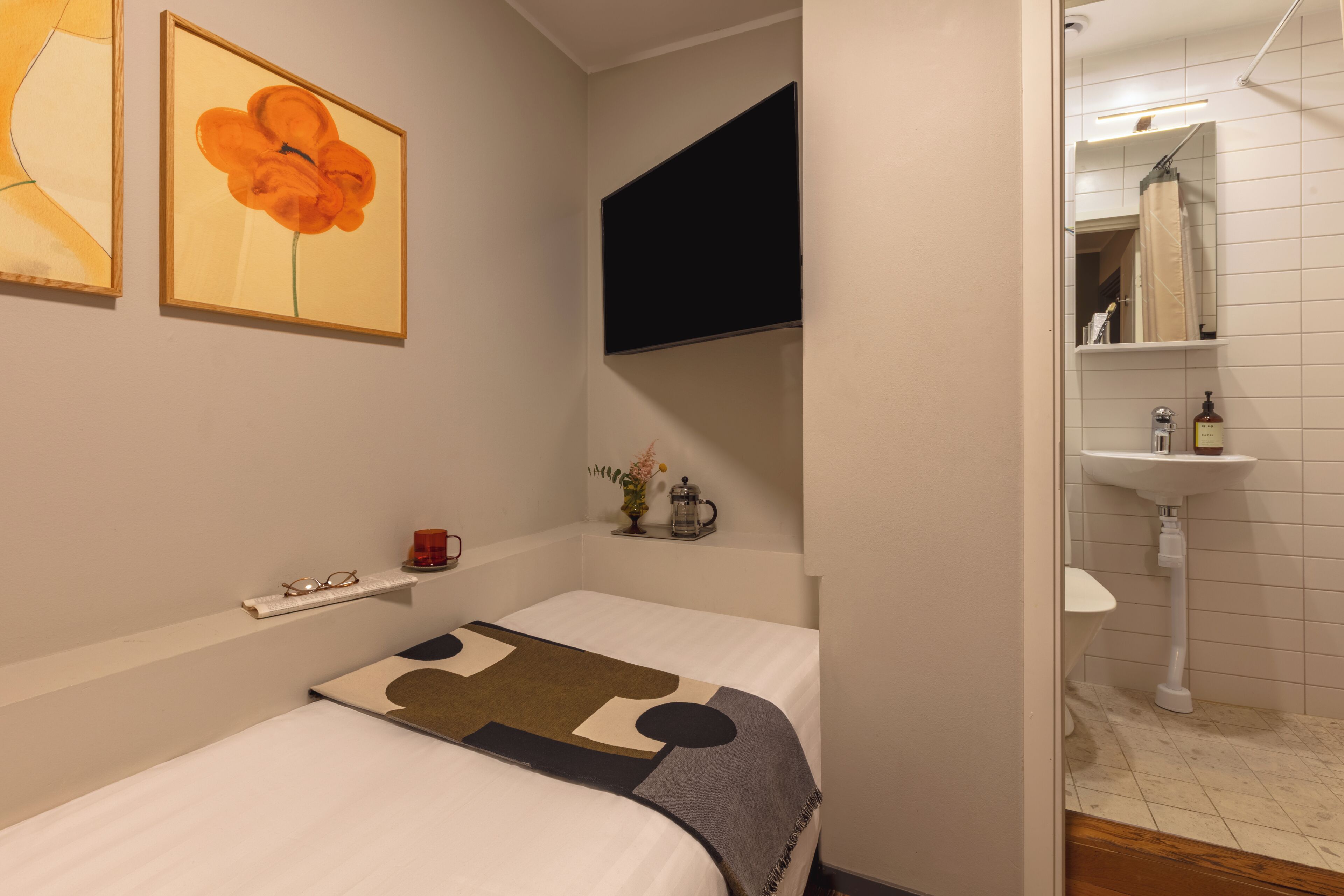 Une chambre à coucher avec un lit, un téléviseur fixé au mur, une petite étagère avec quelques objets, et une salle de bain visible à travers une porte ouverte.