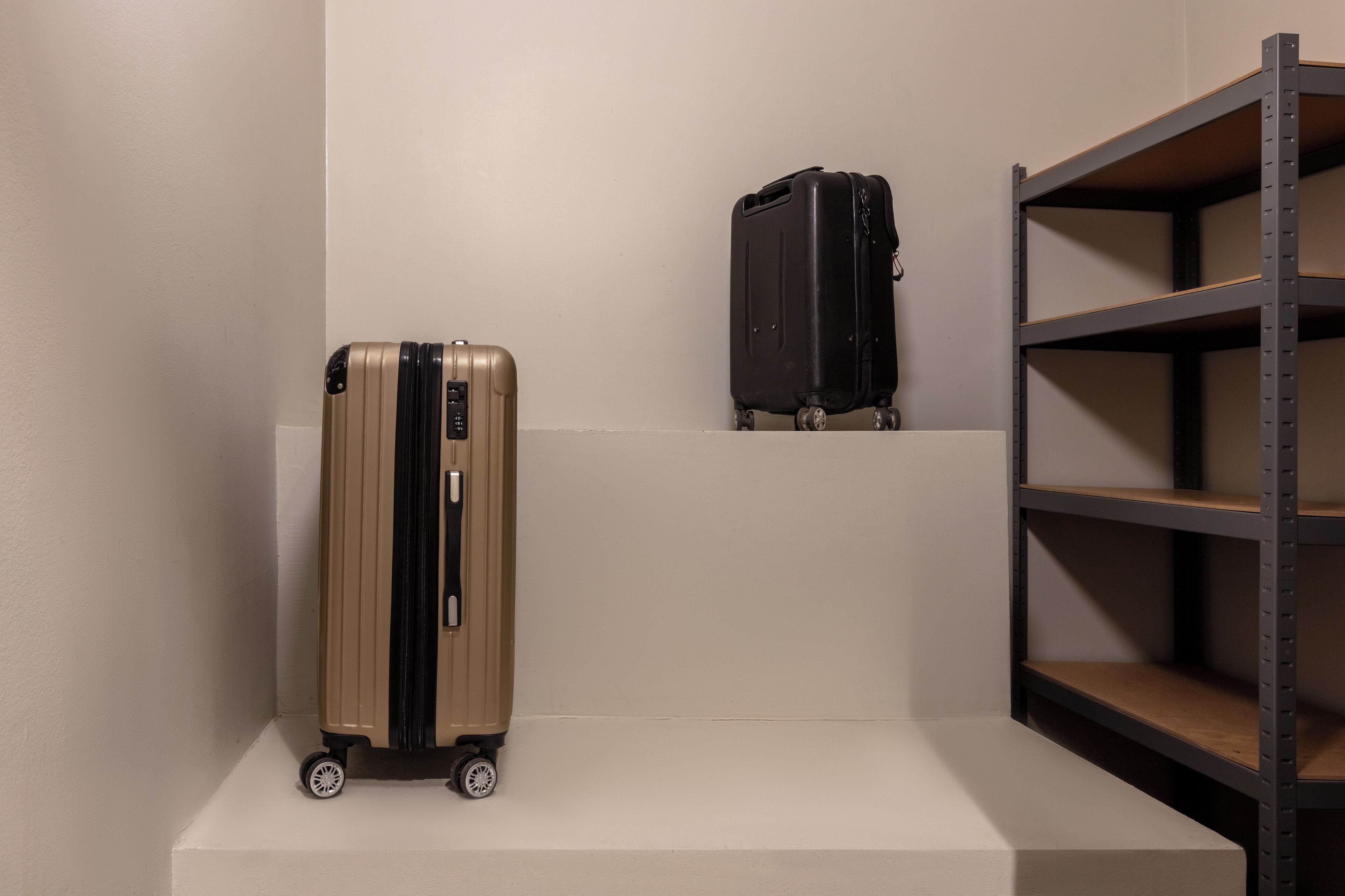 Une valise beige et une valise noire sont posées sur une étagère blanche, devant un mur uni.