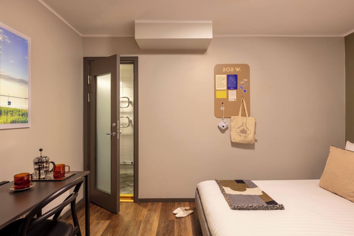 Une chambre d’hôtel comprenant un lit, un bureau avec une machine à café, un tableau d’affichage fixé au mur et une porte donnant accès à une salle de bain.