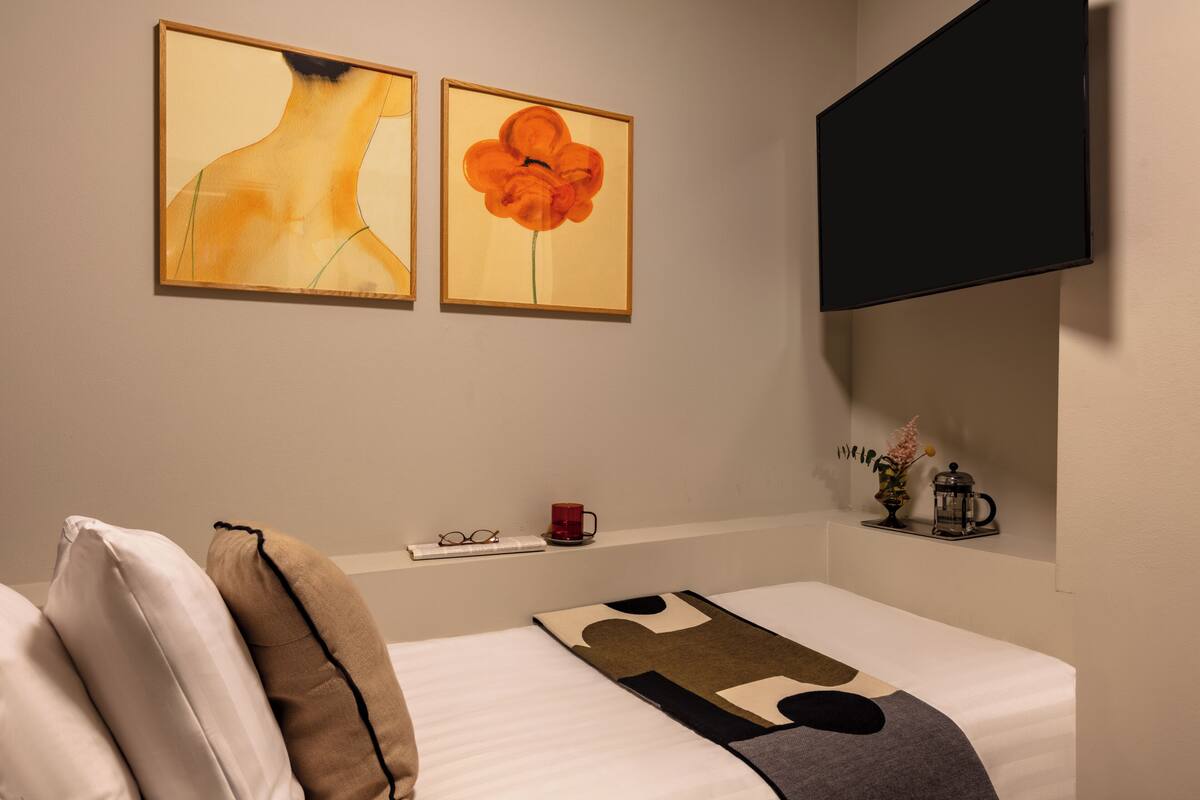Une chambre d’hôtel avec un lit, des oreillers, une table de chevet sur laquelle se trouvent une tasse, des verres et un vase. Deux œuvres d’art encadrées ornent le mur.