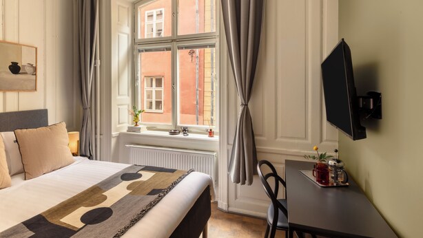 Habitación de hotel con cama, ventana con cortinas, escritorio con cafetera y televisión montada en la pared.