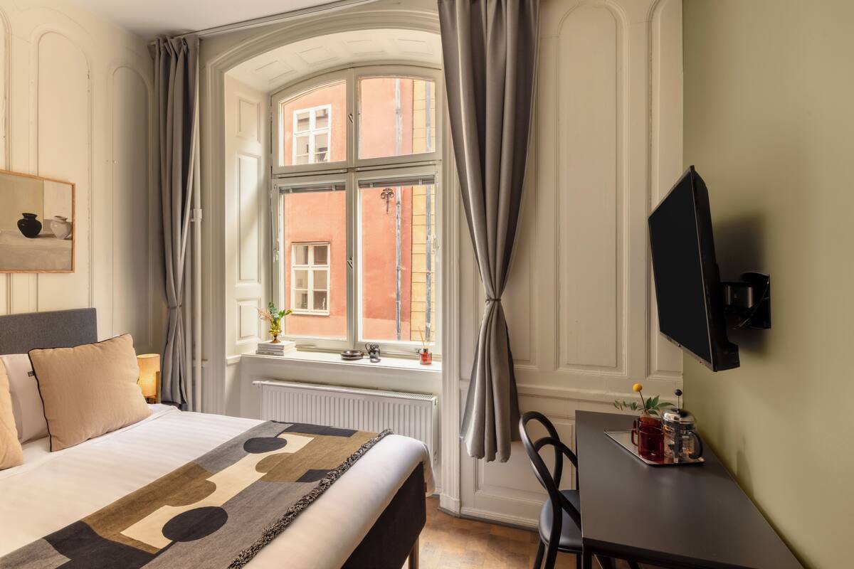 Une chambre d’hôtel avec un lit, une fenêtre avec des rideaux, un bureau avec une machine à café et un téléviseur fixé au mur.