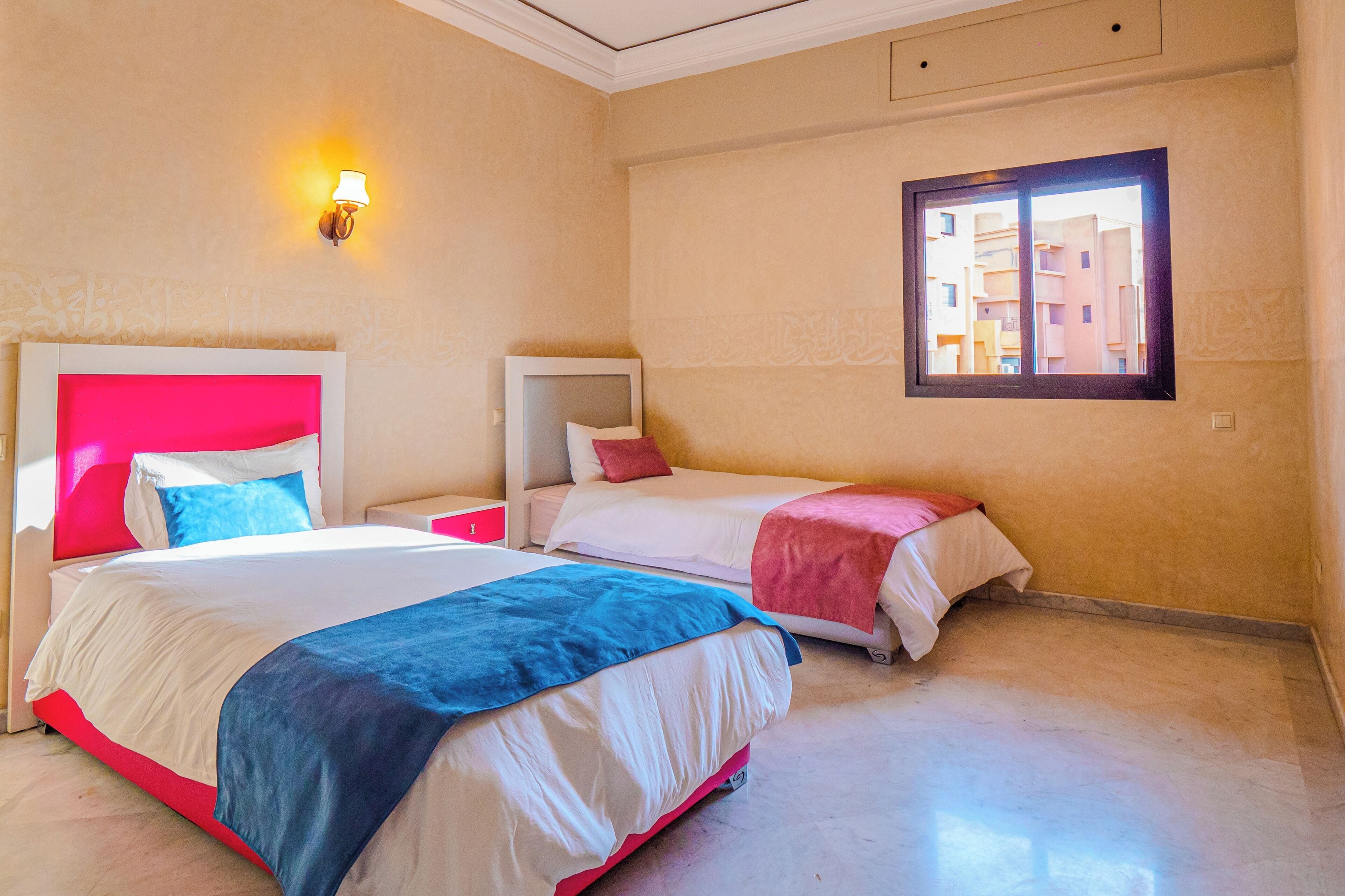 A hotel room with two beds, a nightstand, a wall-mounted TV, and a window with a view of buildings.