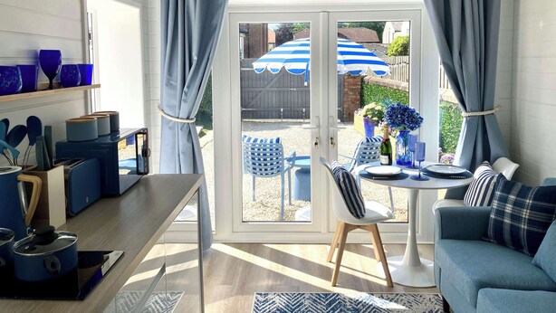 Ein gemütliches Zimmer mit Esstisch, Stühlen und einem blauen Sofa. Ein Fenster mit Vorhängen bietet einen Blick nach draußen.