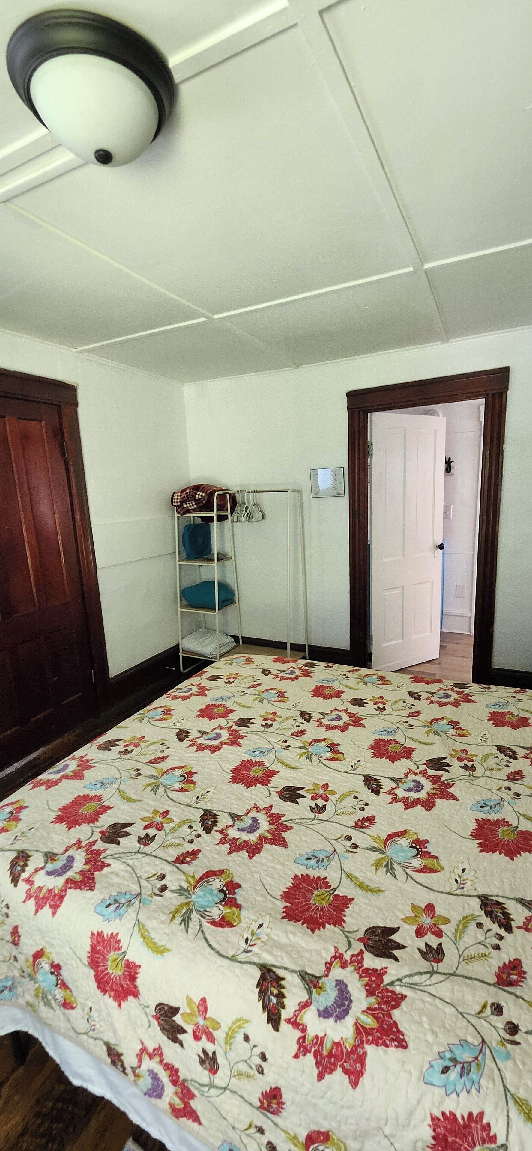 A room with a floral-patterned bedspread, a white ceiling with a light fixture, a metal shelf with items, and a wooden door.