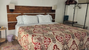 A bedroom with a wooden headboard, a patterned bedspread, a small table with a vase, and a standing fan. - Watkins Glen Overlook Apt (Watkins Glen)