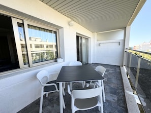 Un balcón con mesa y sillas, un ventanal corredera y vistas a edificios.