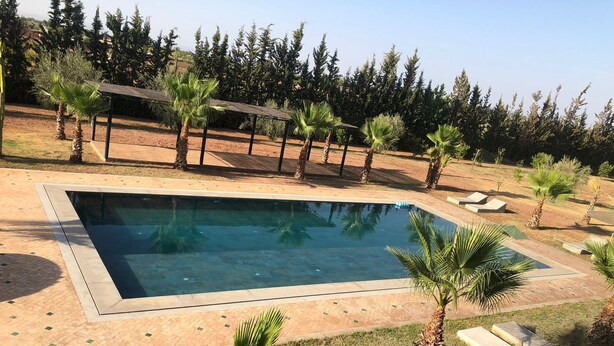 A rectangular swimming pool with a tiled deck surrounding it, palm trees, and a pergola structure. - HOUSE ROUTE DE L'OURIKA MARRAKECH (Marrakech)