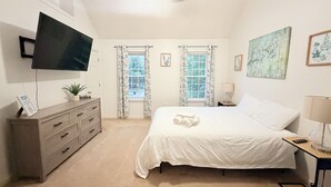 Une chambre à coucher comprenant un lit, un téléviseur, une commode, deux fenêtres avec des rideaux et un ventilateur de plafond.