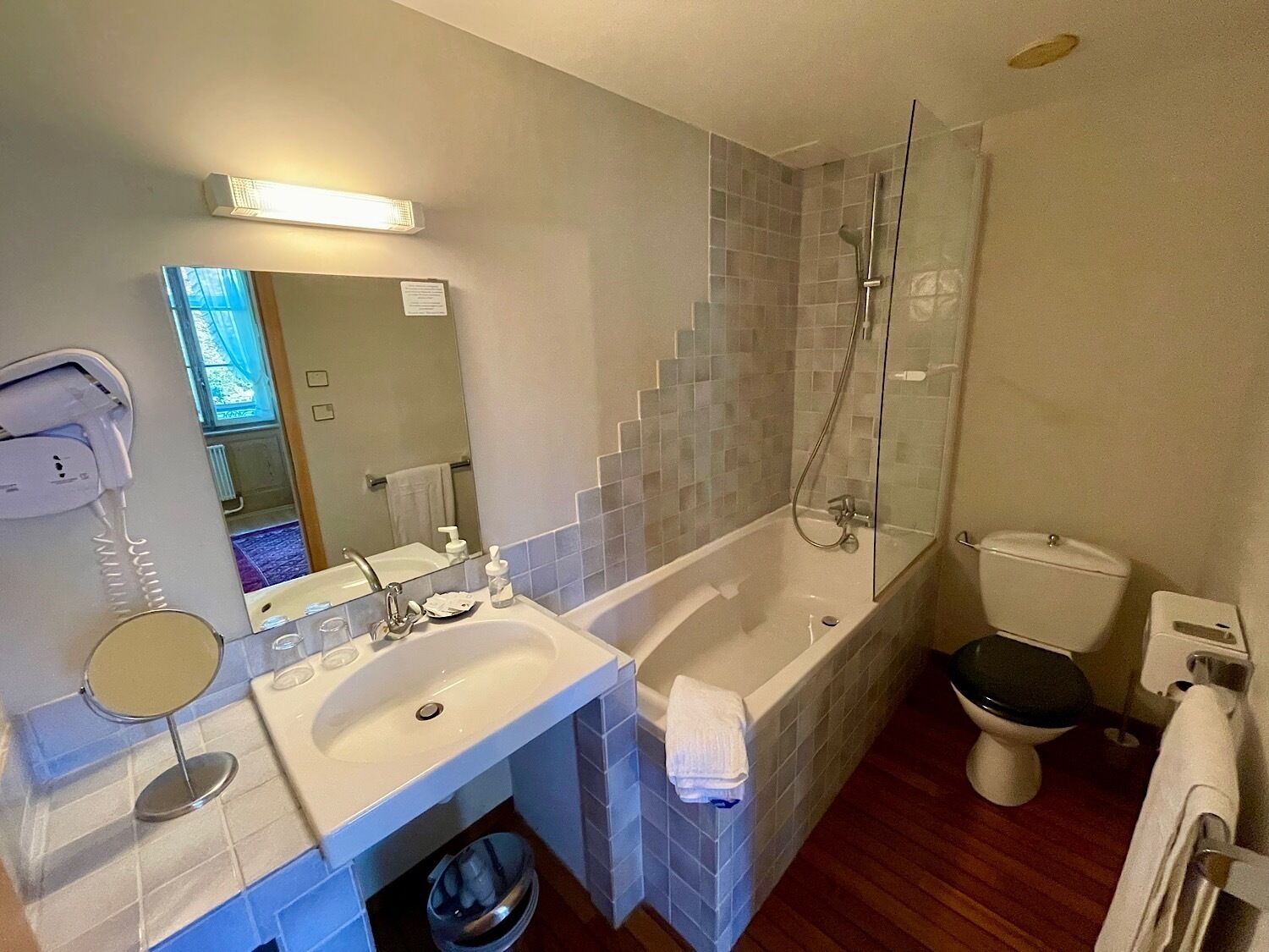 A bathroom with a sink, mirror, handheld showerhead, toilet, and a trash bin.