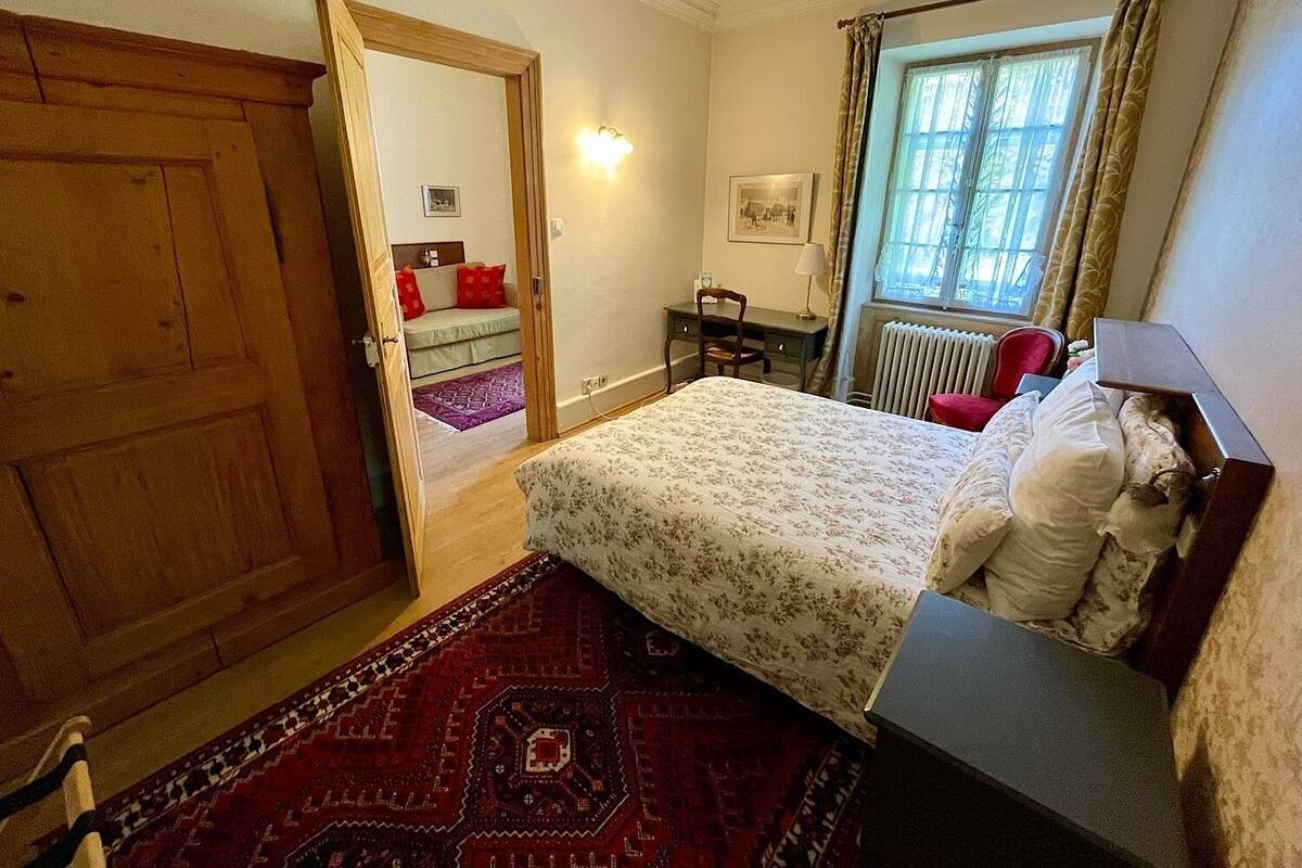 Ein Schlafzimmer mit Bett, Schreibtisch, Stuhl, Heizkörper und einem Fenster mit Vorhängen.