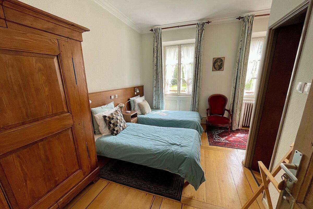 Ein Zimmer mit zwei Betten, einem Holzschrank, einem roten Sessel und Blick ins Freie durch ein mit Vorhängen versehenes Fenster.