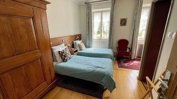 A room with two beds, a wooden wardrobe, a red chair, and a view of the outdoors through a window with curtains.
