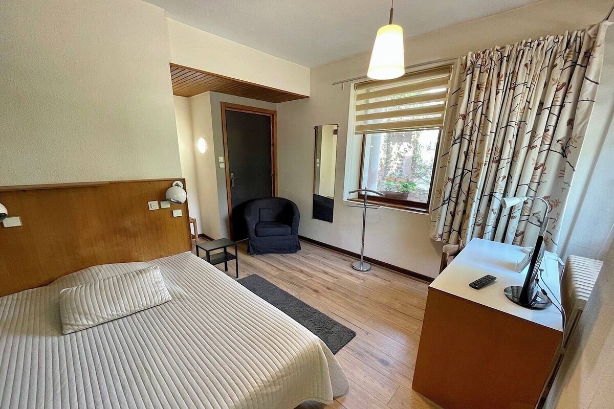 Ein Hotelzimmer mit Bett, Schreibtisch mit Computer, Stuhl, Fenster mit Vorhängen und Tür.