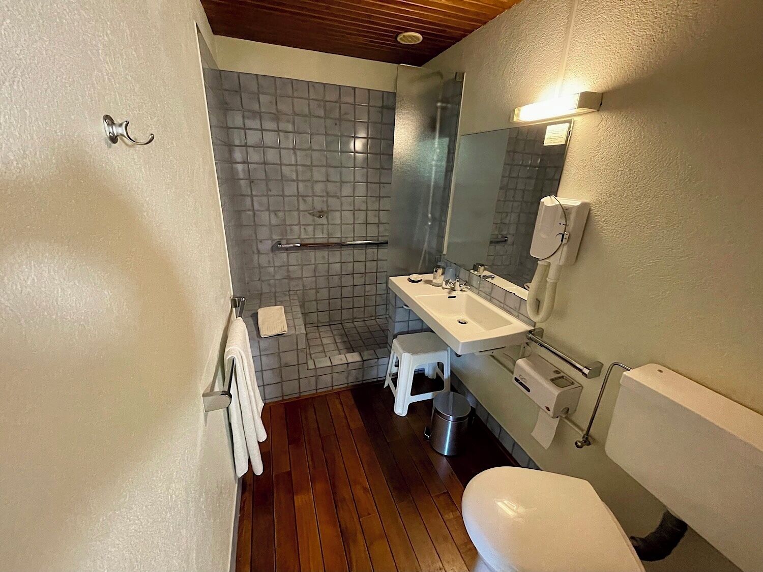 A bathroom with a wooden floor, a white sink, a mirror, a toilet, and a shower area.