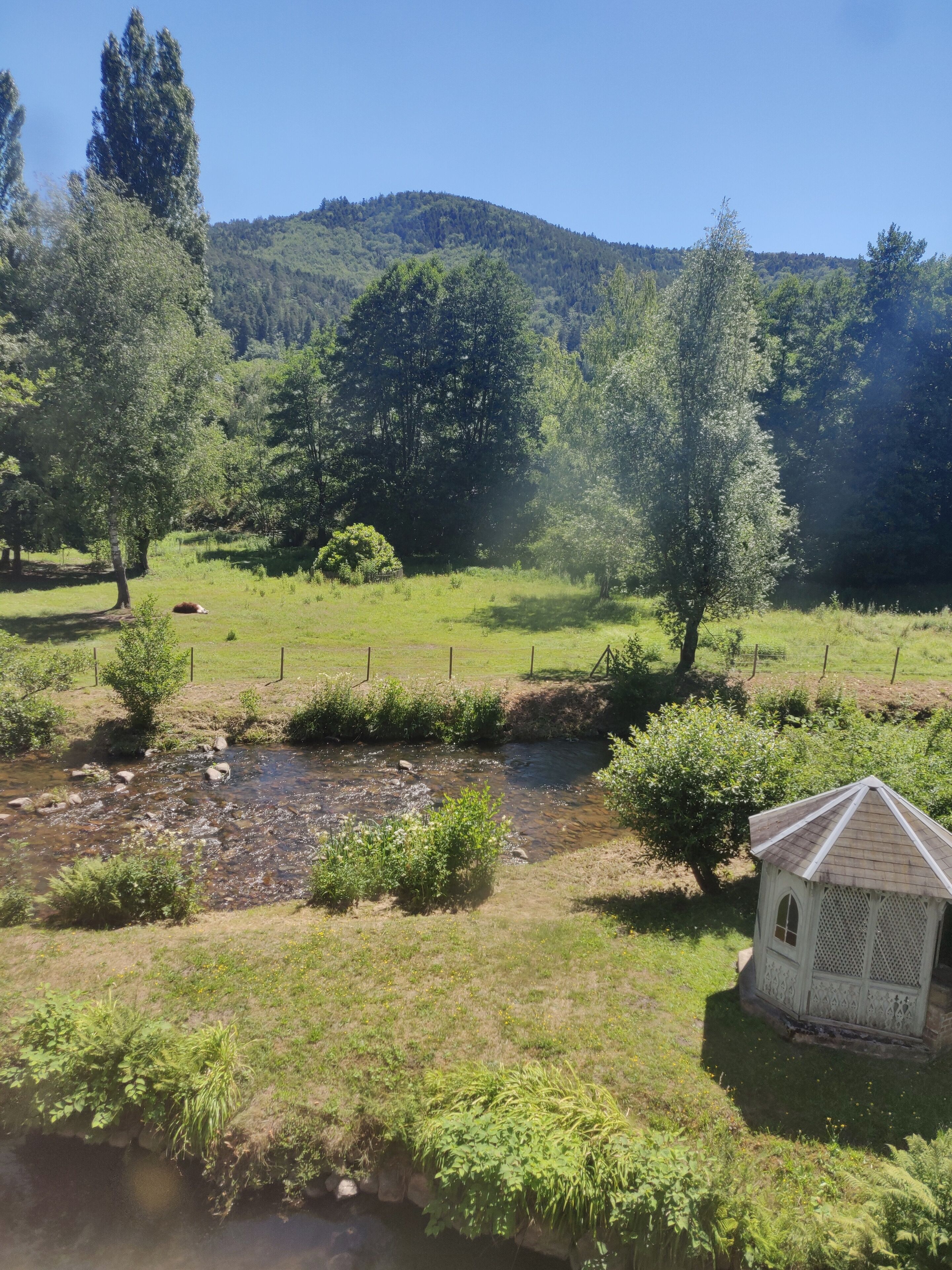 A rural landscape with a small white gazebo, a stream, and a forested hill in the background.