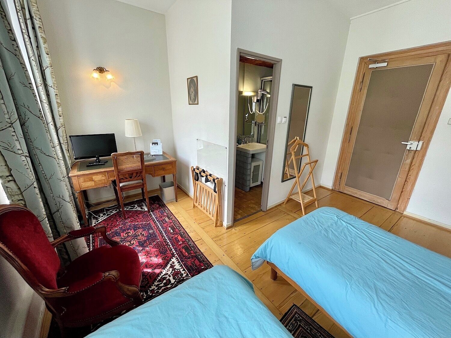A hotel room with two single beds, a desk with a computer, a chair, a red armchair, a mirror, and a bathroom visible through an open door.