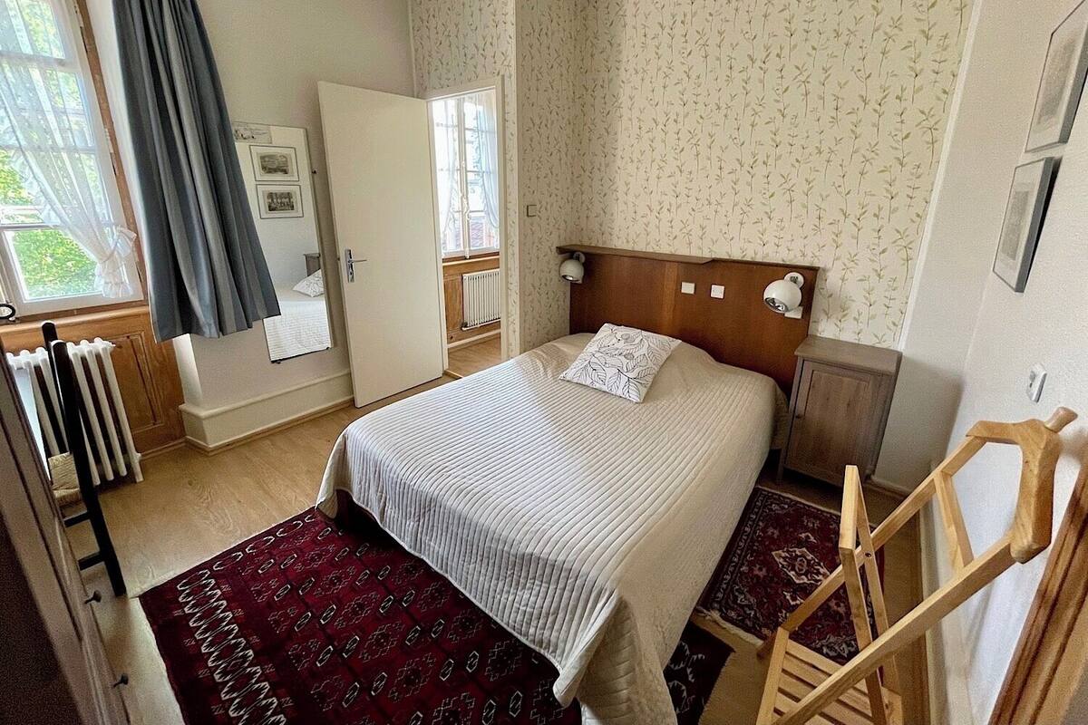 Ein Schlafzimmer mit Bett, Holz-Kopfteil, Nachttisch, Heizkörper, Fenster mit Vorhängen und einem Holzstuhl.