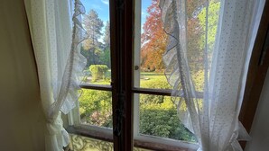 Une fenêtre avec des rideaux blancs donnant sur un jardin planté d’arbres et d’arbustes.