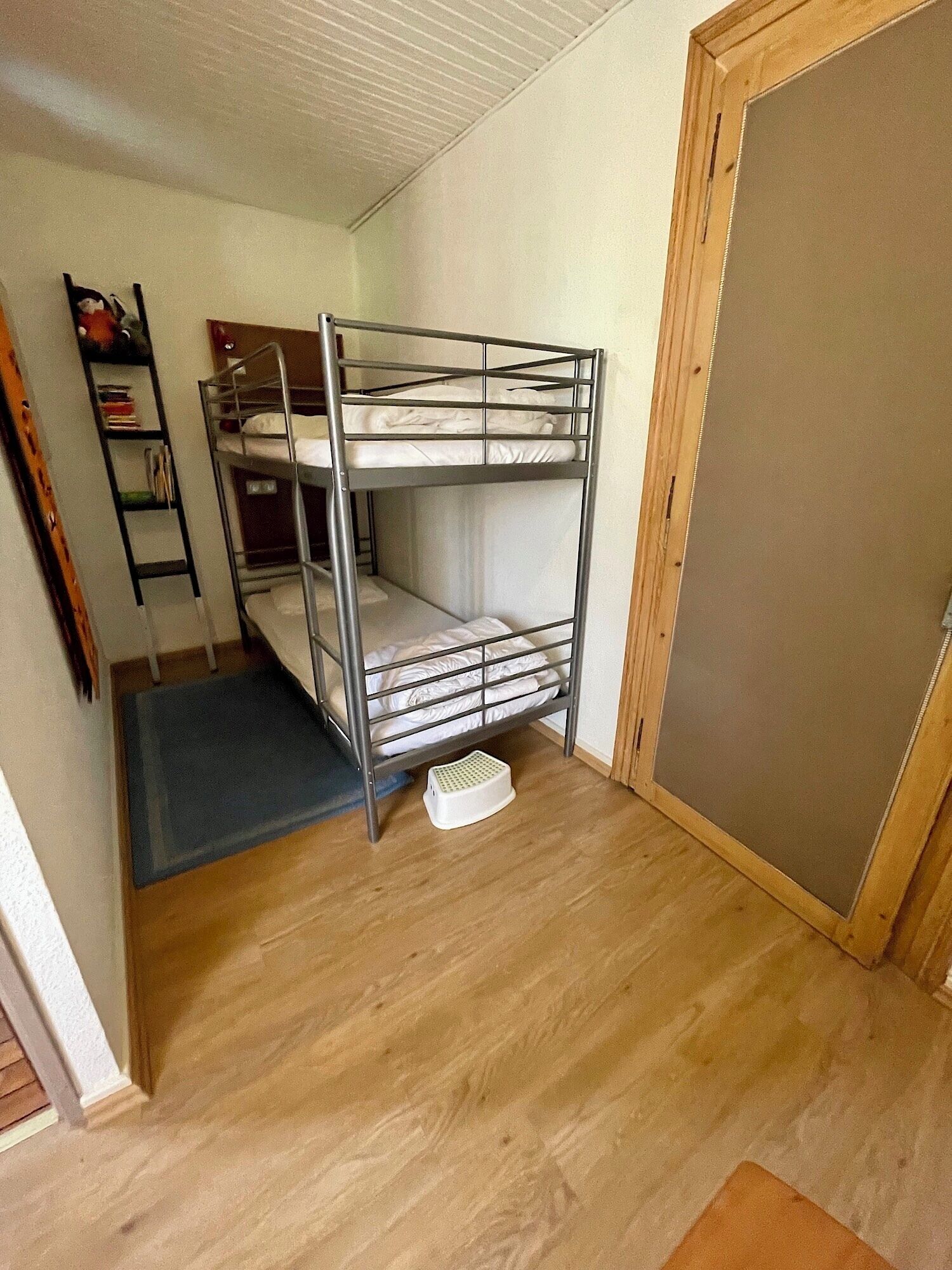 Habitación con literas, suelo de madera, un taburete pequeño y una puerta.