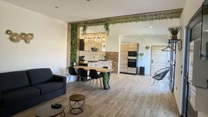 A modern living room with a dark sofa, a coffee table, and a dining area with a wooden table and chairs. - Gîte nature M&J (Calais)