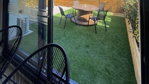 A modern room with large windows, a round wooden table with chairs, and a view of a backyard with artificial grass and a wooden fence. - Gîte nature M&J (Calais)