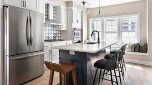 A modern kitchen with a stainless steel refrigerator, white cabinets, a large island with bar stools, and pendant lighting. - Cute and Quaint on Carrollton | Patio & Game Room (Indianapolis)