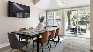A dining area with a large table, chairs, a flat-screen TV, and a view of the backyard through glass doors. - Cute and Quaint on Carrollton | Patio & Game Room (Indianapolis)