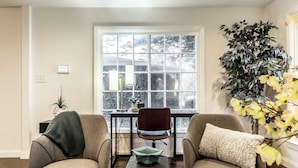 A room with two armchairs, a small table, a window with a view of trees, and a large plant. - The Crestview Bungalow | Steps to Broad Ripple! (Indianapolis)