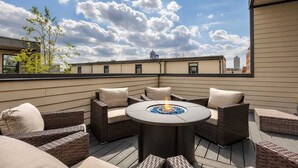 A rooftop patio with wicker furniture, a central fire feature, and a cityscape view.