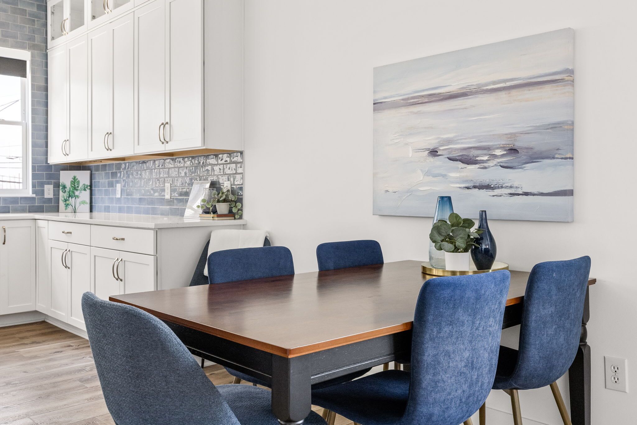 Un comedor con mesa de madera y sillas azules, armarios blancos y un cuadro en la pared.