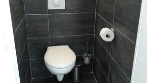 Un piccolo bagno rivestito di piastrelle scure, con il water bianco, un cestino e un portasciugamani.