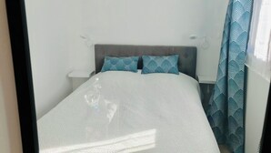 Un letto rifatto con cura, con una coperta a motivi, due cuscini a rombi, una testiera grigia rivestita in tessuto e una finestra con tende a fantasia.