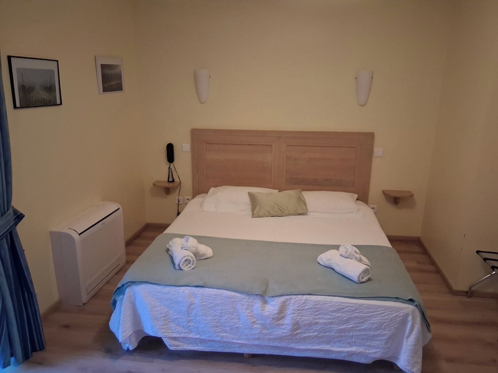 Una cama bien hecha con dos toallas enrolladas, una mesita de noche con lámpara y un teléfono instalado en la pared.