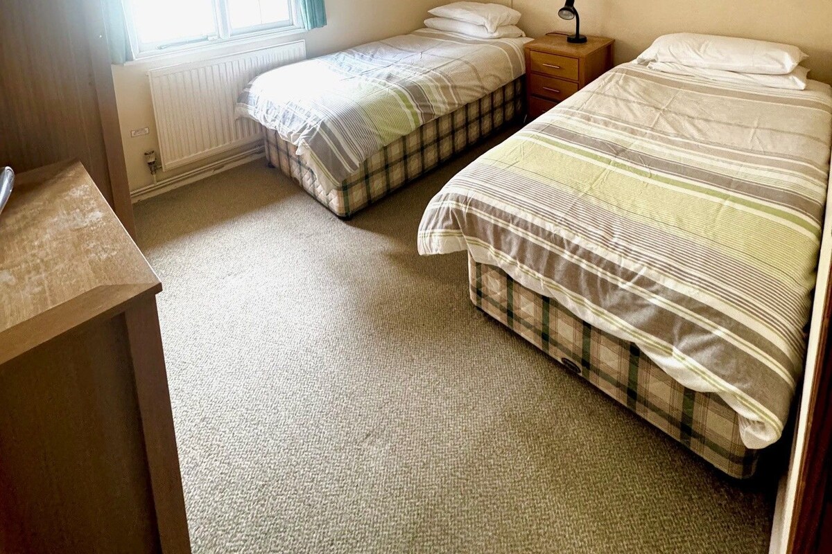 A small, well-lit room with two beds, a radiator, a window with curtains, and a corkboard on the wall.