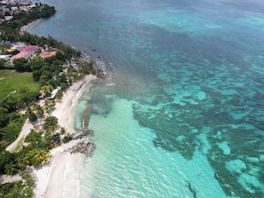 Vista aérea de una zona costera con playa, aguas turquesas cristalinas y un área residencial con casas y vegetación.