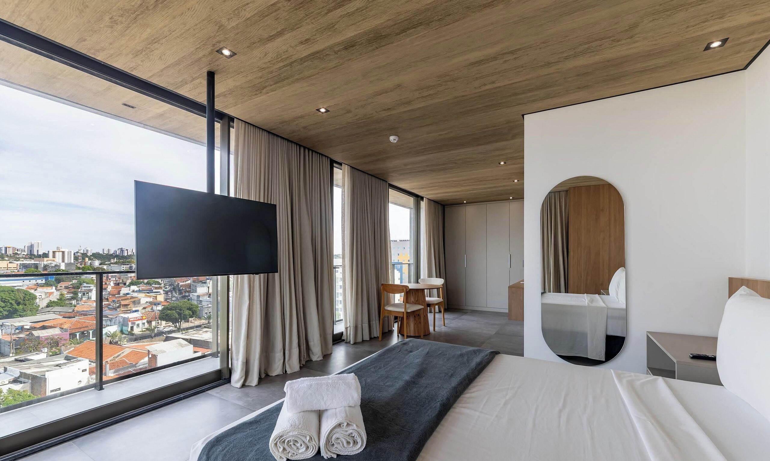 Ein modernes Hotelzimmer mit einem großen Bett, einem Flachbildfernseher, Blick auf die Stadt und einem Essbereich.