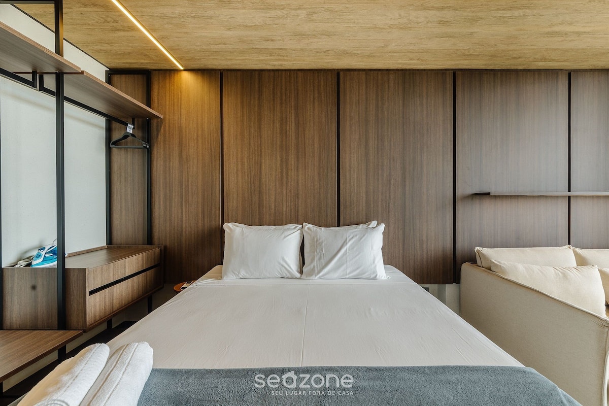 A modern hotel room with a large bed, wooden panel walls, a built-in bench, and a hanging rack.
