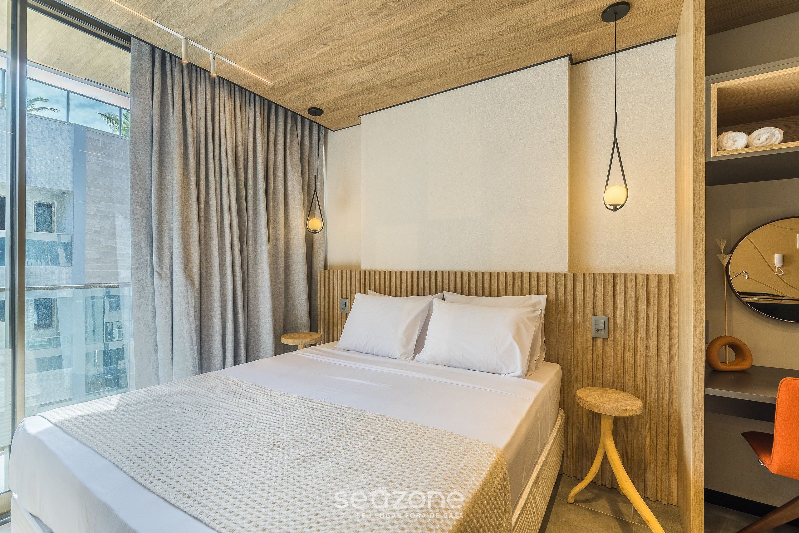 A modern hotel room with a large bed, wooden headboard, a small wooden stool, a round mirror, and a view of the city.