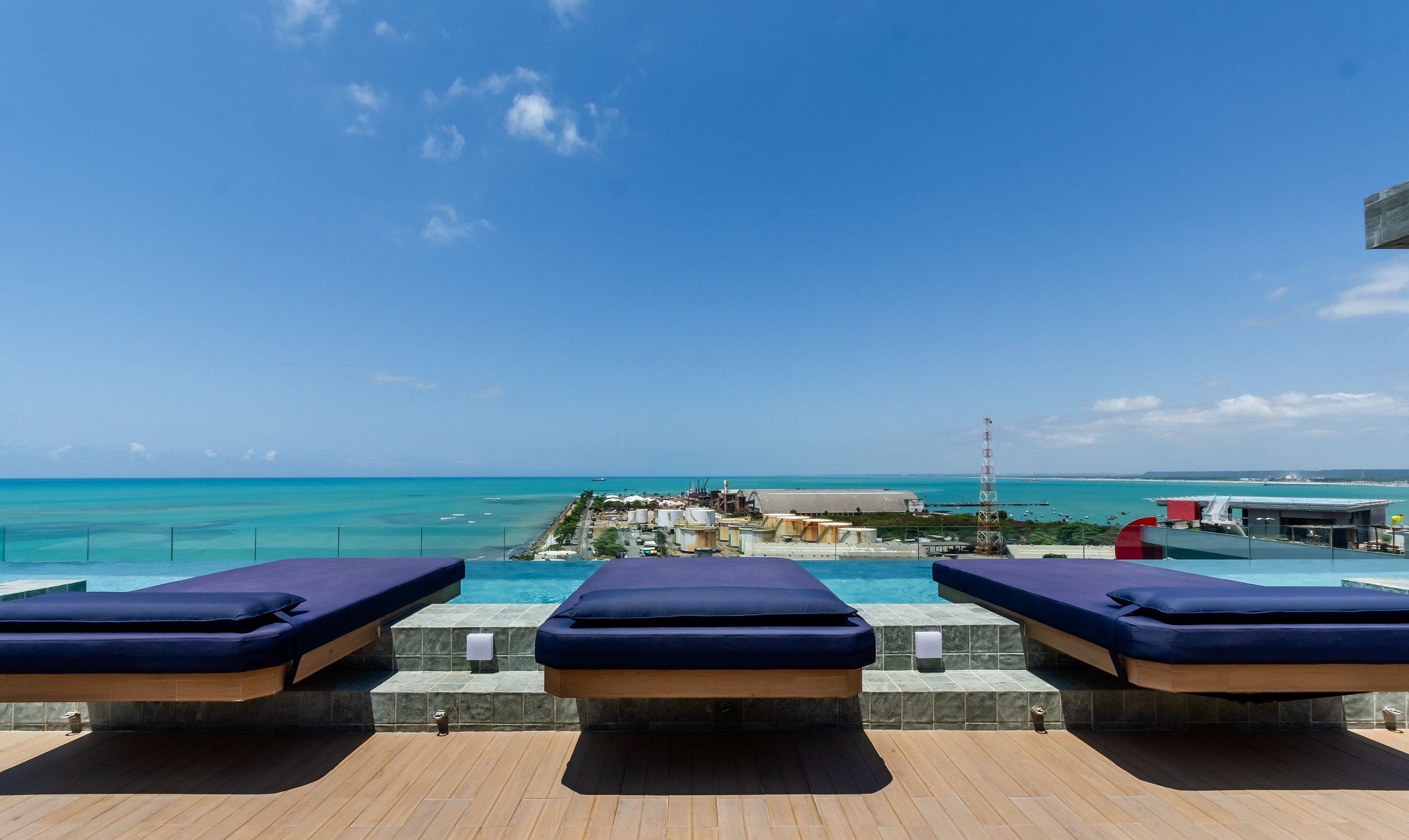 Ein Poolbereich mit Liegestühlen und Blick auf das Meer sowie entfernte Gebäude.