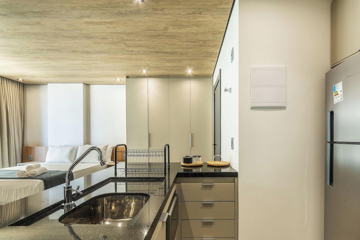 A hotel room with a kitchenette, a bed with rolled towels, a bathroom with a shower, and a ceiling with recessed lighting.