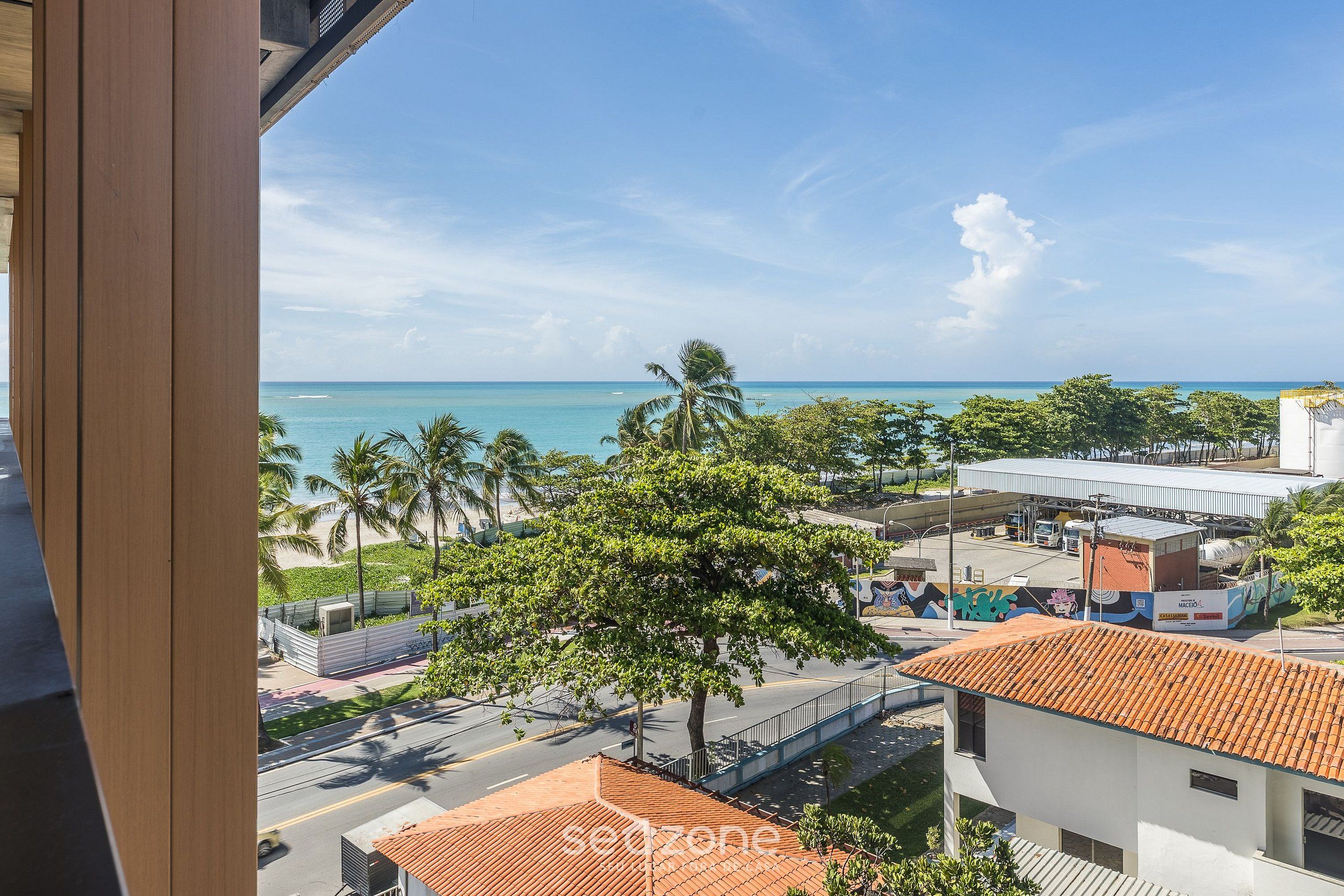 A coastal view with a clear sky, palm trees, and a road leading to a building with a tiled roof.