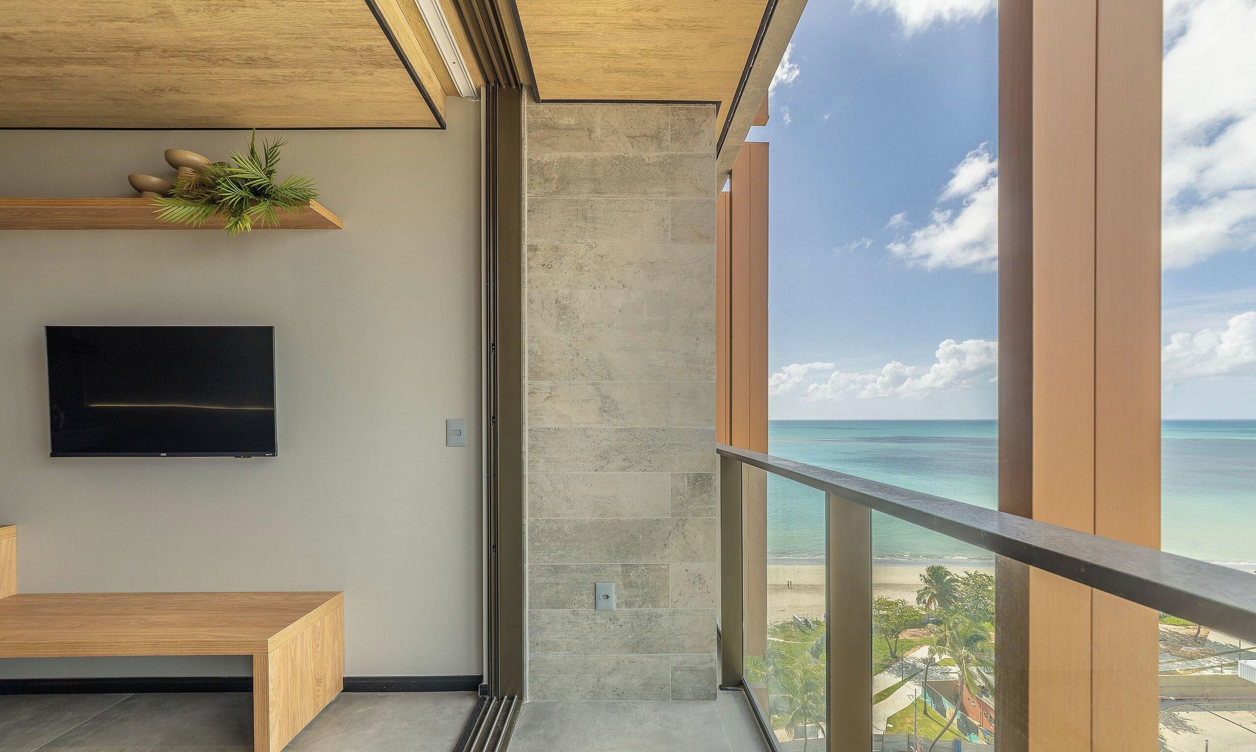 Une chambre moderne dotée d’une grande fenêtre donnant sur une plage, d’un téléviseur à écran plat fixé au mur et d’un banc en bois.