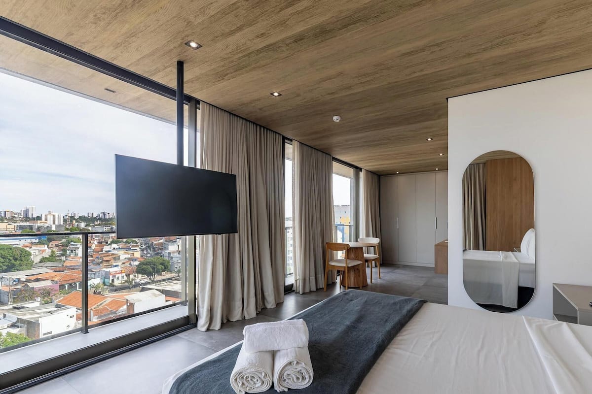 A modern hotel room with a large window, a flat-screen TV, a bed, a desk, and a mirror.