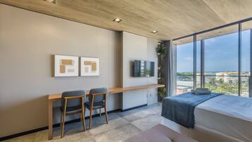 A modern bedroom with a large window, a bed, a desk with chairs, and artwork on the wall.