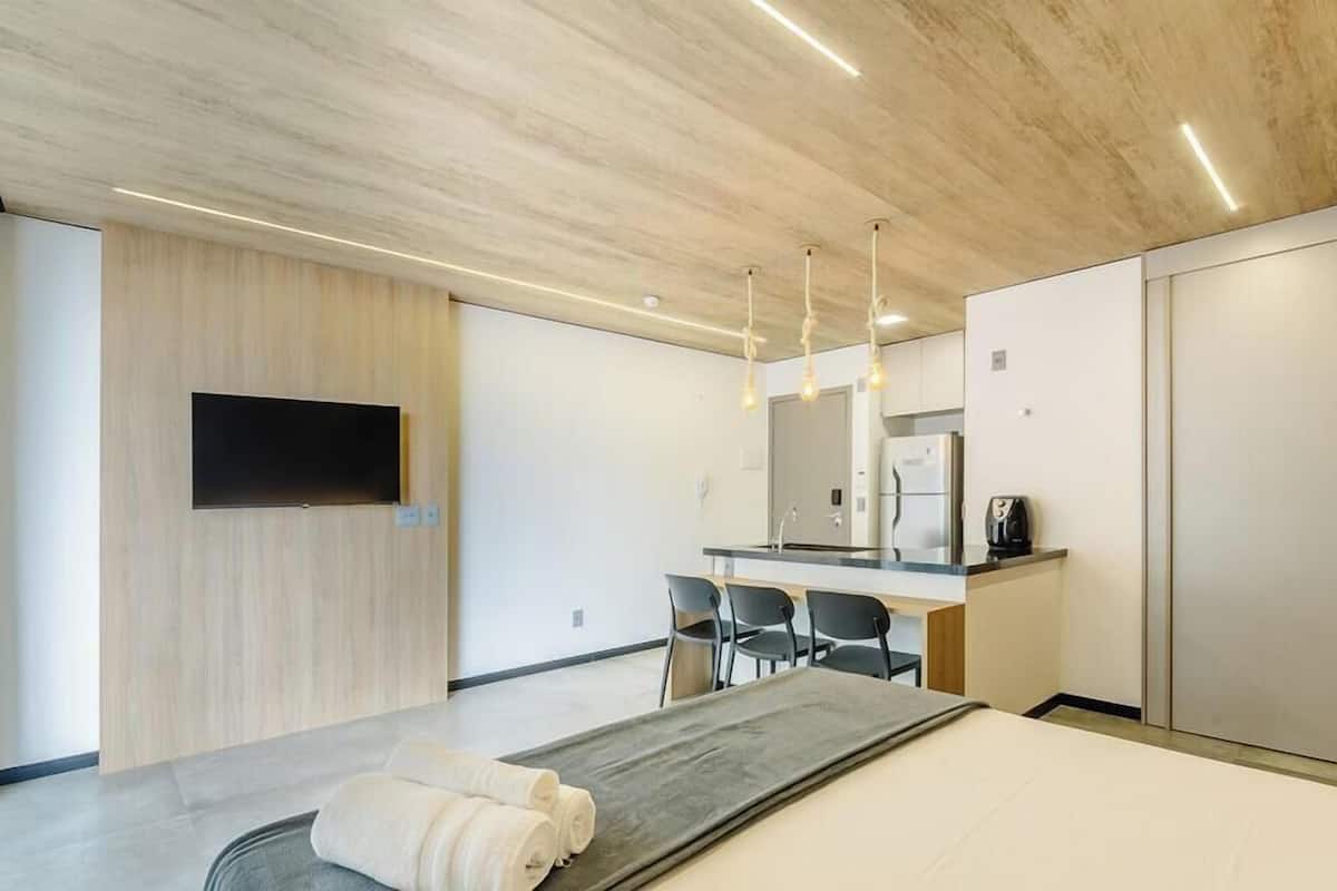 A modern hotel room with a wooden ceiling, a flat-screen TV mounted on the wall, a dining area with chairs, a kitchen area with a refrigerator, and a bed with a gray headboard.