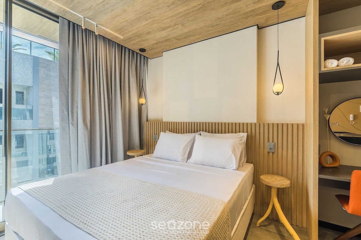 A modern hotel room with a large bed, wooden headboard, a small wooden stool, a round mirror, and a view of the city.