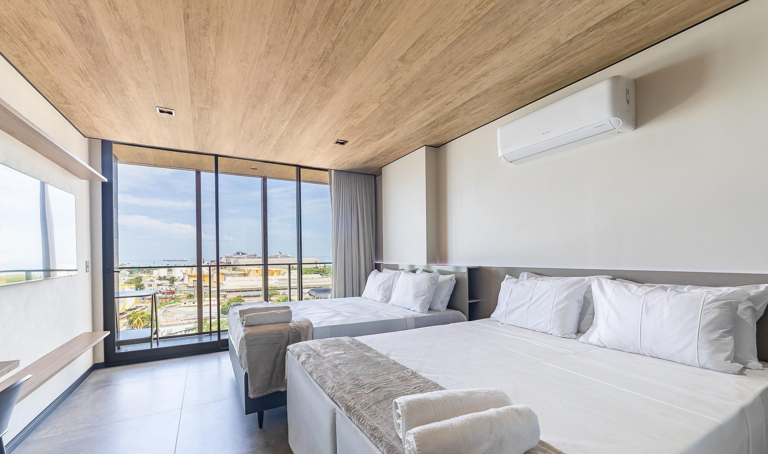 Una habitación de hotel moderna con una cama grande, una cama más pequeña y un sofá. Hay una ventana amplia con vista a la ciudad y al mar.
