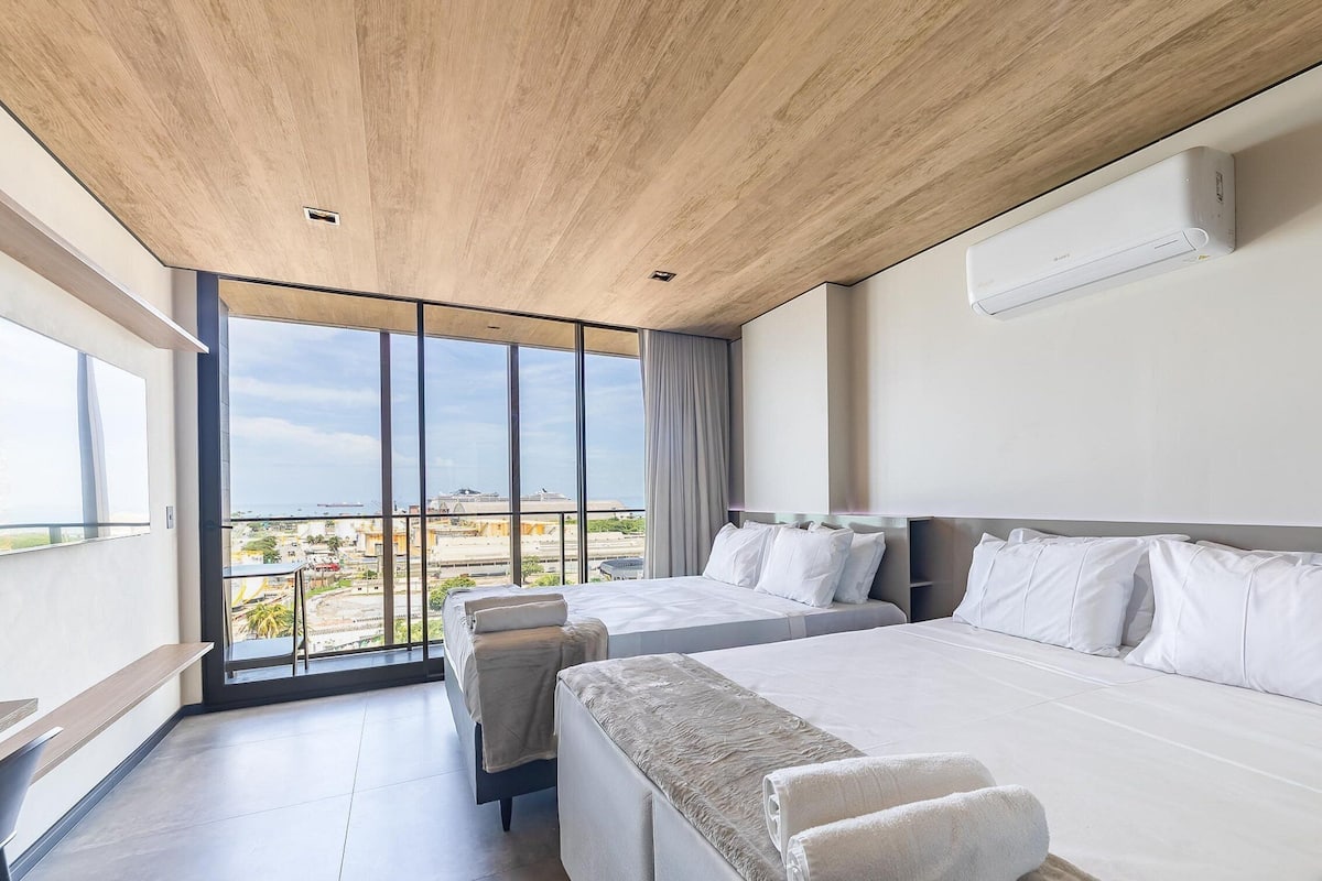 A modern hotel room with a large bed, a smaller bed, and a sofa. There is a large window with a view of the city and the sea.