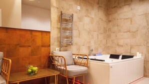 A bathroom with a bathtub, a towel rack, a chair, and a table with a fruit bowl.